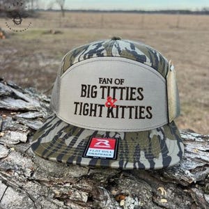 May include: A flat bill snapback hat with a camouflage pattern on the bill and a tan front panel. The front panel features the text "FAN OF BIG TITTIES & TIGHT KITTIES" in black lettering. A small tag is attached to the side.