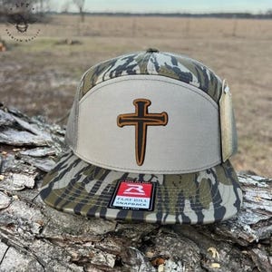 May include: A camouflage flat bill snapback hat with a brown cross design on a light tan patch. The hat features a mesh back and a tag. The brim has a camouflage pattern of green, brown, and black.