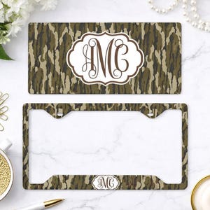 May include: A license plate and frame set with a camouflage pattern in shades of brown and green. The license plate features a white oval with the monogram "MG" in brown script. The license plate frame has the same camouflage pattern and a smaller monogram.
