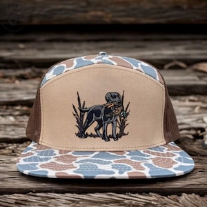 May include: A tan and brown trucker hat with a camouflage pattern on the brim. The hat features a black Labrador Retriever carrying a duck in its mouth, embroidered on the front panel. The hat has a brown mesh back.