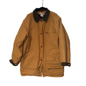 Puede incluir: Chaqueta L.L.Bean de color marrón claro con cuello y puños verde oscuro. La chaqueta tiene cuatro bolsillos delanteros, cierre de botones y un pequeño logotipo en el bolsillo del pecho. Cuelga de una percha negra.
