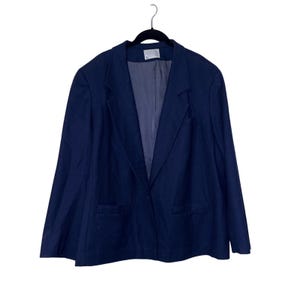 Puede incluir: Un blazer azul marino con solapa de muesca, dos bolsillos delanteros y cierre de un solo botón. El blazer está colgado de una percha negra sobre un fondo blanco. El forro interior es de un tono azul más claro.