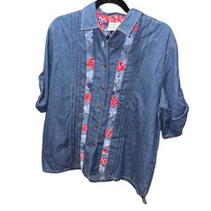 Può includere: Una camicia di jeans blu con bottoni e maniche corte. La camicia presenta pannelli decorativi sul davanti con tessuto a fantasia rosso e blu. Il colletto e la fodera interna hanno un motivo floreale rosso. La camicia è appesa a una gruccia nera.