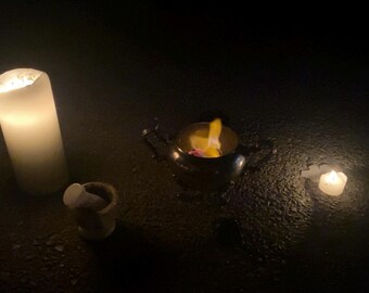 Puede incluir: Una escena oscura con tres fuentes de luz. Una vela grande y blanca está encendida, junto con una vela pequeña y redonda y un cuenco con una llama. La escena es oscura, con las llamas proporcionando la única luz.