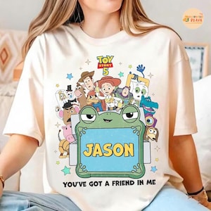 May include: Cream-colored t-shirt featuring a colorful graphic of Toy Story 5 characters. A green frog holds a sign with the name "JASON" in yellow. The text "YOU'VE GOT A FRIEND IN ME" is printed at the bottom.