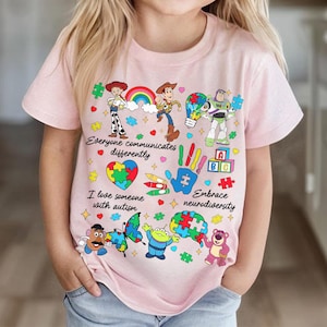 May include: Pink t-shirt featuring Toy Story characters and puzzle pieces. The shirt includes the phrases "Everyone communicates differently", "I love someone with autism", and "Embrace neurodiversity". The design incorporates a rainbow and various colorful elements.