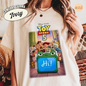 May include: Ivory-colored t-shirt featuring a graphic of the Disney Pixar movie "Toy Story 5." The graphic includes characters like Woody and Buzz Lightyear, with a green frog-shaped frame that says "Hi! Let's play!"