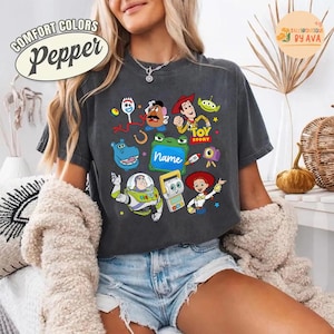 May include: Dark gray Comfort Colors Pepper t-shirt featuring various Toy Story characters. The shirt includes images of Woody, Buzz Lightyear, and other characters from the movie. The text "TOY STORY" is also visible.