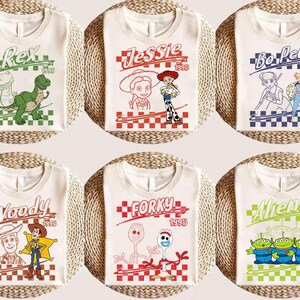 May include: Six white t-shirts featuring characters from the movie Toy Story. Each shirt has a different character design, including Rex, Jessie, Bo Peep, Woody, Forky, and Aliens, with the year 1995. The designs are set against a checkered background.