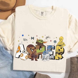 May include: Cream-colored t-shirt featuring the name "Asher" in a Star Wars-themed design. The letters are decorated with characters like BB-8, Chewbacca, R2-D2, C-3PO, and Grogu. The shirt has a crew neck and short sleeves.