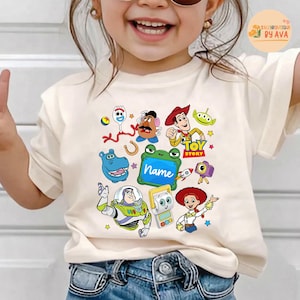 May include: Cream-colored t-shirt featuring colorful cartoon characters from the Toy Story movie. The design includes Woody, Buzz Lightyear, and other characters, along with the text "Toy Story" and a space for a name.