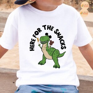 May include: White t-shirt with a green cartoon dinosaur holding a snack, and the text "HERE FOR THE SNACKS" in a black arch. The dinosaur has black mouse ears. A fun and playful design for children.