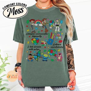 May include: A moss green t-shirt featuring a colorful graphic of Toy Story characters, puzzle pieces, and the phrases "Everyone communicates differently," "I love someone with autism," and "Embrace neurodiversity." The shirt has a Comfort Colors brand label.