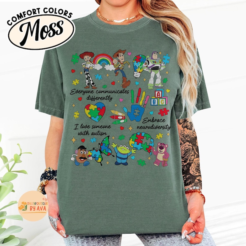 May include: A moss green t-shirt featuring a colorful graphic of Toy Story characters, puzzle pieces, and the phrases "Everyone communicates differently," "I love someone with autism," and "Embrace neurodiversity." The shirt has a Comfort Colors brand label.
