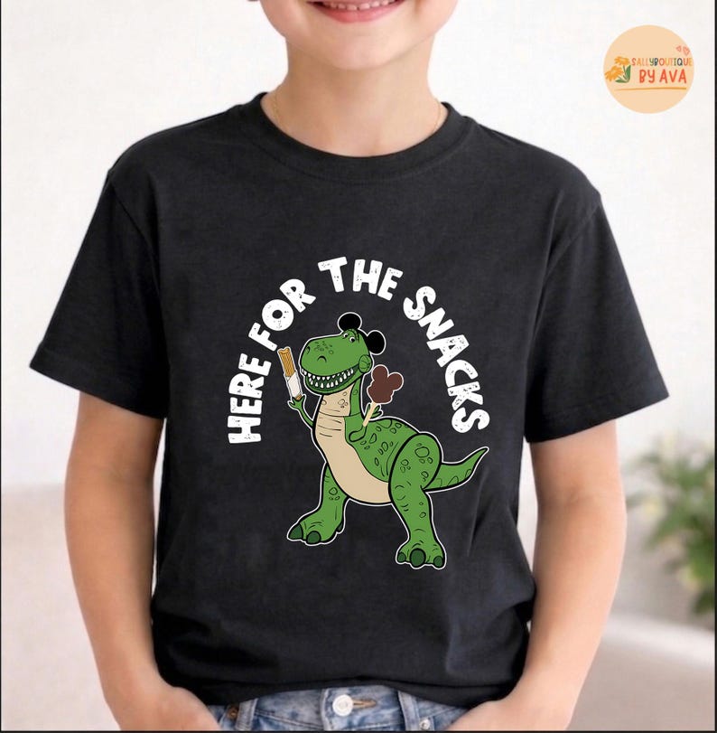 May include: Black t-shirt featuring a green cartoon dinosaur holding snacks. The text "HERE FOR THE SNACKS" is in a white, circular font. The dinosaur has a white belly and is wearing a black hat.