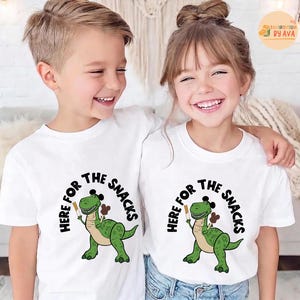 May include: Two children wearing white t-shirts featuring a green dinosaur graphic holding a snack. The text "HERE FOR THE SNACKS" is printed above and around the dinosaur. The children are smiling and embracing each other.