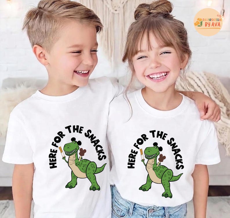 May include: Two children wearing white t-shirts featuring a green dinosaur graphic holding a snack. The text "HERE FOR THE SNACKS" is printed above and around the dinosaur. The children are smiling and embracing each other.