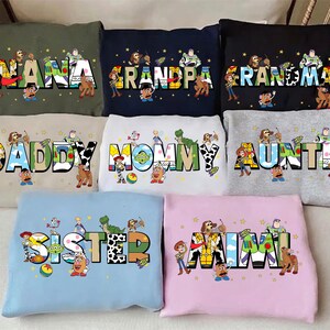 May include: A collection of colorful sweatshirts featuring characters from a popular animated movie. Each sweatshirt displays a different family title, such as "NANA", "GRANDPA", "MOMMY", and "AUNTIE", with the characters integrated into the lettering.