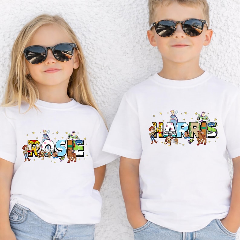 May include: Two children wearing white t-shirts with personalized names in colorful, cartoon-style lettering. The names are decorated with images of characters from a popular animated movie. Both children are wearing sunglasses.