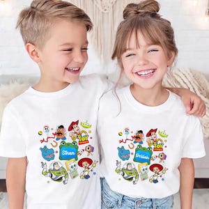 May include: Two children wearing white t-shirts featuring a colorful Toy Story graphic. The shirts display various characters from the movie, including Woody, Buzz Lightyear, and others. The name "Ethan" is on one shirt and "Delilah" on the other.