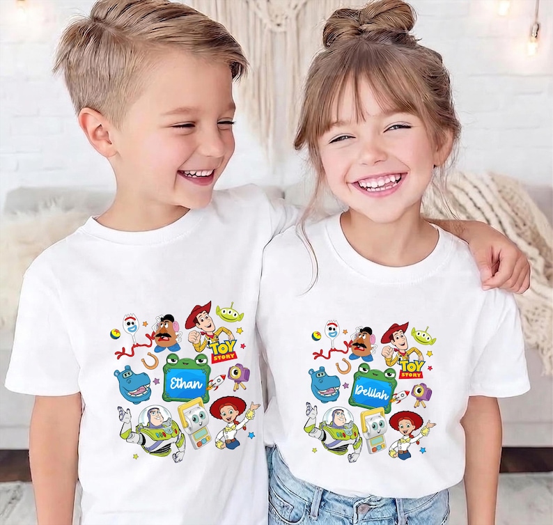 May include: Two children wearing white t-shirts featuring a colorful Toy Story graphic. The shirts display various characters from the movie, including Woody, Buzz Lightyear, and others. The name "Ethan" is on one shirt and "Delilah" on the other.