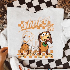 May include: A light gray sweatshirt featuring a cartoon image of Slinky Dog from Toy Story. The design includes the word "Slinky" in orange, the year "1995", and a checkered background. The sweatshirt has a crew neck and long sleeves.