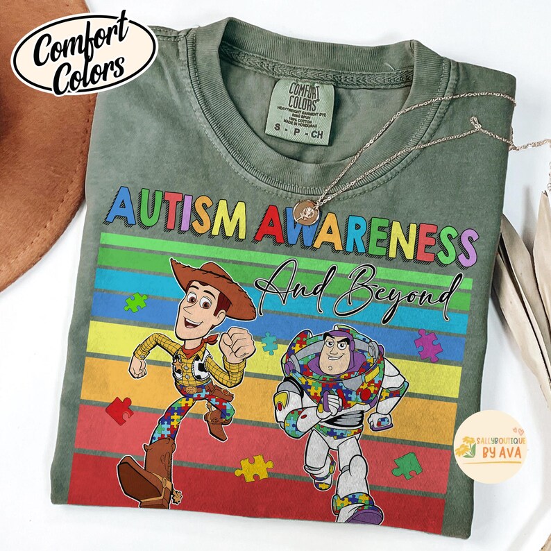 May include: Green Comfort Colors t-shirt with the words "Autism Awareness And Beyond" in colorful letters. The shirt features Woody and Buzz Lightyear characters with puzzle piece designs. A gold necklace is draped over the shirt.