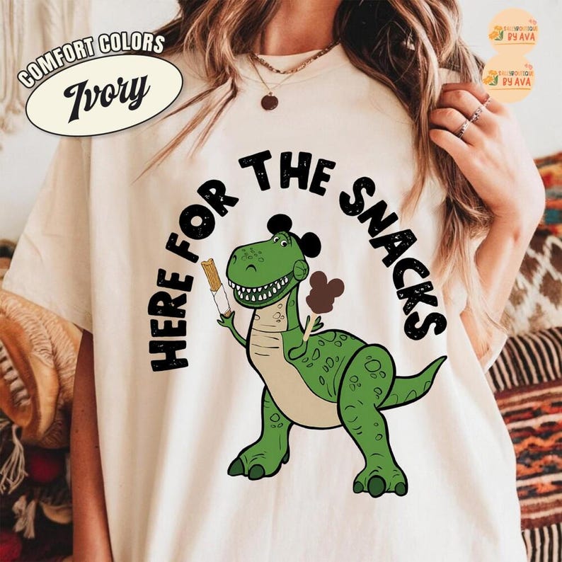 May include: Ivory t-shirt featuring the phrase "HERE FOR THE SNACKS" around a green cartoon dinosaur holding a snack. The shirt also has the words "COMFORT COLORS" and "Ivory" in an oval.