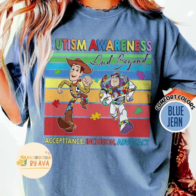May include: Blue jean-colored t-shirt with a colorful graphic promoting autism awareness. The design features Woody and Buzz Lightyear characters, the text "AUTISM AWARENESS And Beyond", and the words "ACCEPTANCE, INCLUSION, ADVOCACY".