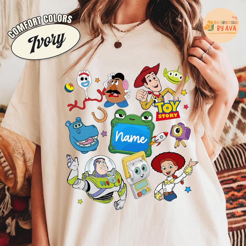 May include: Ivory-colored t-shirt featuring a colorful Toy Story graphic. The design includes cartoon characters like Woody, Buzz Lightyear, and others. The shirt also has the text "Comfort Colors" and "Ivory" in an oval shape.