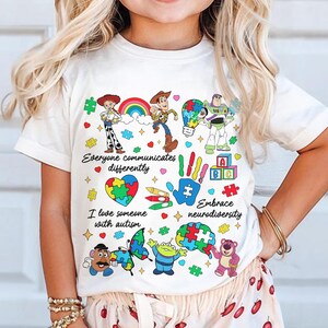 May include: White t-shirt featuring colorful cartoon characters and the text "Everyone communicates differently" and "I love someone with autism." The shirt includes a rainbow, puzzle pieces, and the phrase "Embrace neurodiversity."