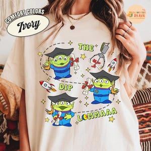 May include: Ivory Comfort Colors t-shirt featuring a cartoon alien design. The design includes aliens in graduation caps, rockets, and the text "THE DIP LOMMAAA". The shirt has a crew neck and short sleeves.
