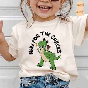 May include: A cream-coloured t-shirt with a green cartoon dinosaur holding snacks. The text "HERE FOR THE SNACKS" is arched above the dinosaur in black. The dinosaur has a friendly expression and is holding a snack in each hand.