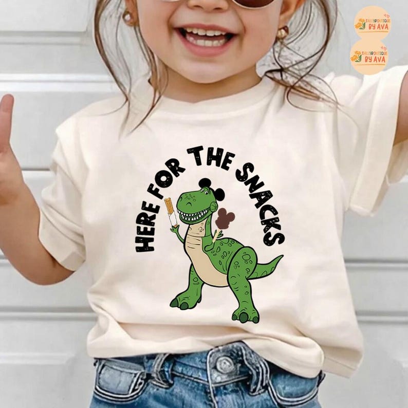 May include: A cream-coloured t-shirt with a green cartoon dinosaur holding snacks. The text "HERE FOR THE SNACKS" is arched above the dinosaur in black. The dinosaur has a friendly expression and is holding a snack in each hand.