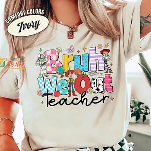 May include: Ivory-colored t-shirt with a colorful graphic that says "Bruh We Out Teacher." The graphic features cartoon characters, flowers, and a globe. The shirt has a crew neck and short sleeves.
