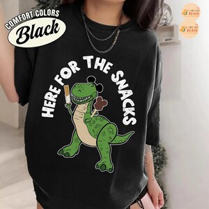 May include: Black t-shirt featuring a green cartoon dinosaur holding a snack and ice cream cone. The text "HERE FOR THE SNACKS" is in white around the dinosaur. The shirt also has the text "COMFORT COLORS Black" in a white oval.