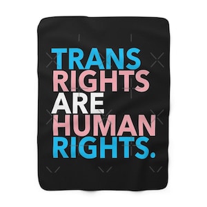 Könnte beinhalten: Eine schwarze Decke mit dem Text "TRANS RIGHTS ARE HUMAN RIGHTS" in Blau, Rosa und Weiß. Die Decke hat ein weiches, plüschiges Aussehen, und der Text ist mittig auf der Vorderseite platziert.