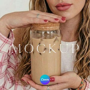 May include: A clear glass jar filled with iced coffee, topped with a cork lid. The word "MOCKUP" is displayed in white across the jar. The Canva logo is visible at the bottom. The jar is held by a person with red nail polish.