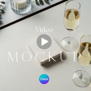 May include: A square white coaster mockup positioned between two tall stemmed glasses filled with a pale yellow liquid. The text Video MOCKUP is centered over the coaster, and a small circular Canva logo is visible at the bottom center.