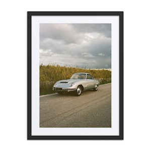 May include: A framed print featuring a silver vintage sports car on a paved road. The car is parked on the side of the road with tall grass and a cloudy sky in the background. The car has a sleek, aerodynamic design.