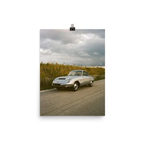 May include: A silver vintage sports car is parked on a paved road, with tall grass and a cloudy sky in the background. The car has a sleek, aerodynamic design and is captured in a scenic outdoor setting. The image is printed on a poster.