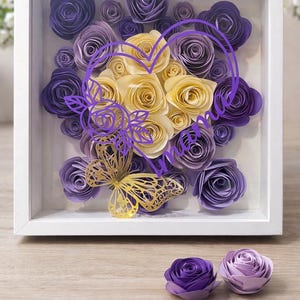 Könnte beinhalten: Handgefertigte Muttertags-Schattenbox mit Papierrosen in Lila- und Gelbtönen. Ein goldener Schmetterling und ein lila herzförmiges Design sind ebenfalls enthalten. Die Schattenbox befindet sich in einem weißen Rahmen.