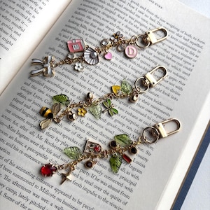 May include: Two gold-coloured keychains with decorative charms, including flowers, leaves, and a bee. The charms are in various colours, including red, green, and yellow. The keychains are on an open book.