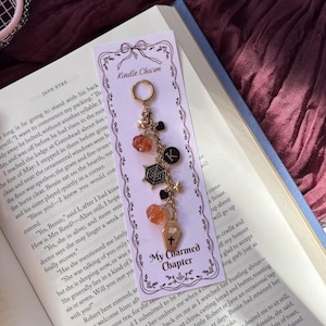 May include: A gold-toned metal charm accessory attached to a light purple card labelled Kindle Charm and My Charmed Chapter. The charm features a gold hoop, small stars, black hearts, orange pumpkin beads, a black spiderweb, a letter K, and a coffin.