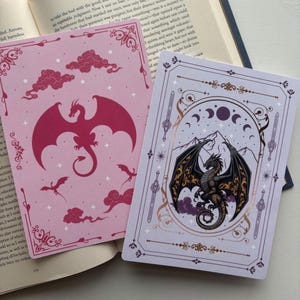 May include: Two fantasy-themed cards. One is pink with a red dragon and cloud designs. The other is lavender with a black dragon, mountain, and moon phases. Both cards have decorative borders and are open on a book.