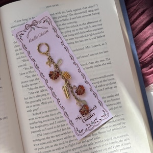 May include: A gold-toned metal charm for an e-reader featuring a hoop attachment and a chain adorned with miniature pendants including brown maple leaves, a pinecone, and a coffee cup, displayed on a light purple card labelled Kindle Charm.