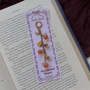 May include: A gold-toned metal charm accessory featuring a small hoop and a chain with dangling charms including a crescent moon, a book, an acorn, a pinecone, and several orange beads, attached to a light purple card labelled Kindle Charm.