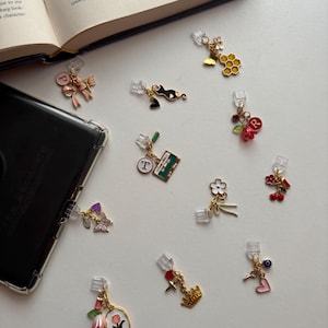 May include: A collection of decorative phone charms with various designs, including a pink bow, a black cat, a cassette tape, a flower, cherries, a crown, and a heart. Each charm has a clear plastic clip for attachment.