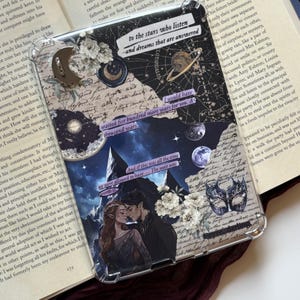 May include: A clear protective tablet case featuring a collage of celestial and fantasy imagery, including a crescent moon, a planet with rings, white flowers, a masquerade mask, and an illustration of two figures, with text reading to the stars who listen and dreams that are answered.