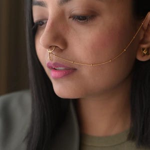 May include: A gold nose ring featuring a small cross pendant and a delicate chain that connects to the ear. The jewellery is worn by a person with dark hair and a green top.